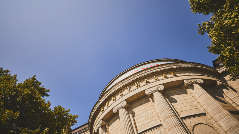 Altbau der Hamburger Kunsthalle von schräg unten fotografiert, halbrunder Gebäudeteil aus Sandstein mit Säulen und Verzierungen, oben die Andeutung eines Kuppeldachs.