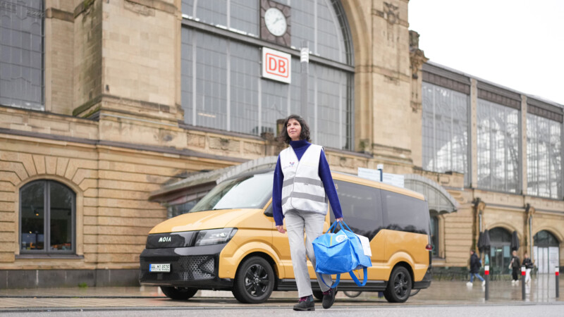 Frau mit Packtasche steht vor einem On-Demand-Shuttle von Moia, das vor dem Hamburger Bahnhof Dammtor parkt