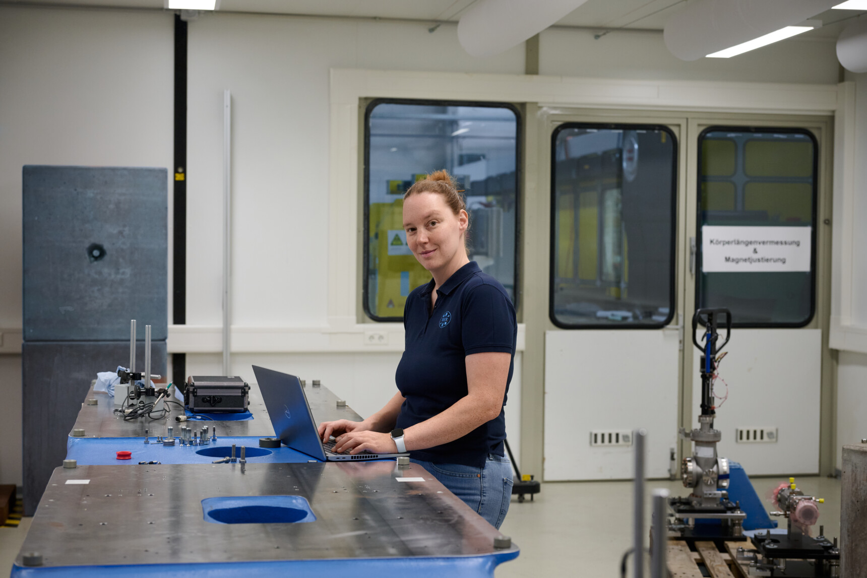 Woman in navy polo shirt working on laptop at industrial workstation with tools and equipment.