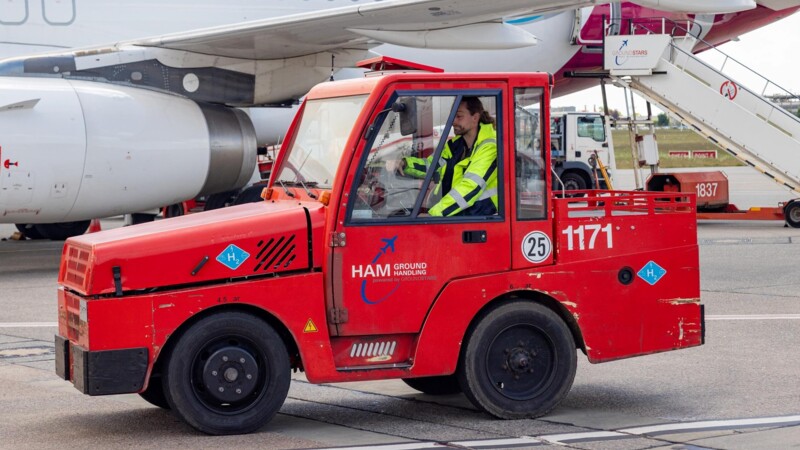 Roter Wasserstoff Gepäckschlepper vor einem Flugzeug am Hamburg Airport. Das Fahrzeug trägt einen Aufkleber mit der Aufschrift "H2". Ein Mitarbeiter mit Warnjacke fährt den Gepäckschlepper