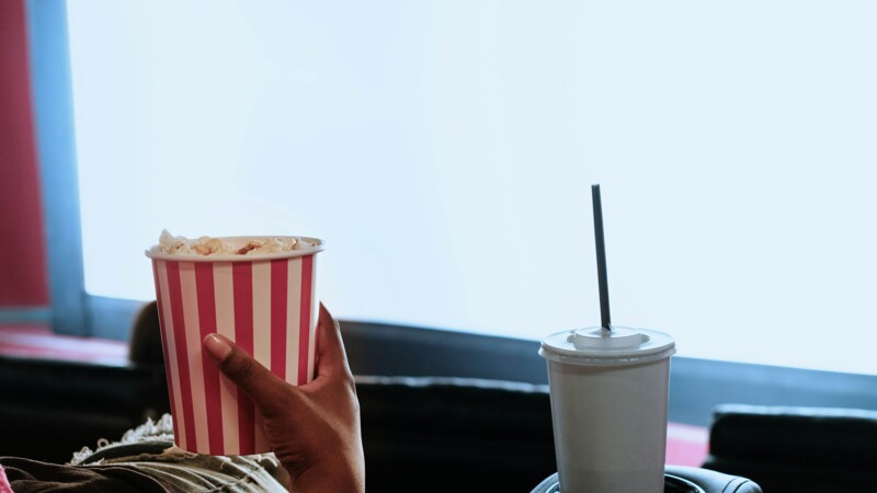 Frau im Kino, die einen Becher Popcorn in der Hand hält, daneben ein Becher mit Strohhalm, im Vordergrund sieht man die Kinoleinwand, Symbolbild Kino-Kongress