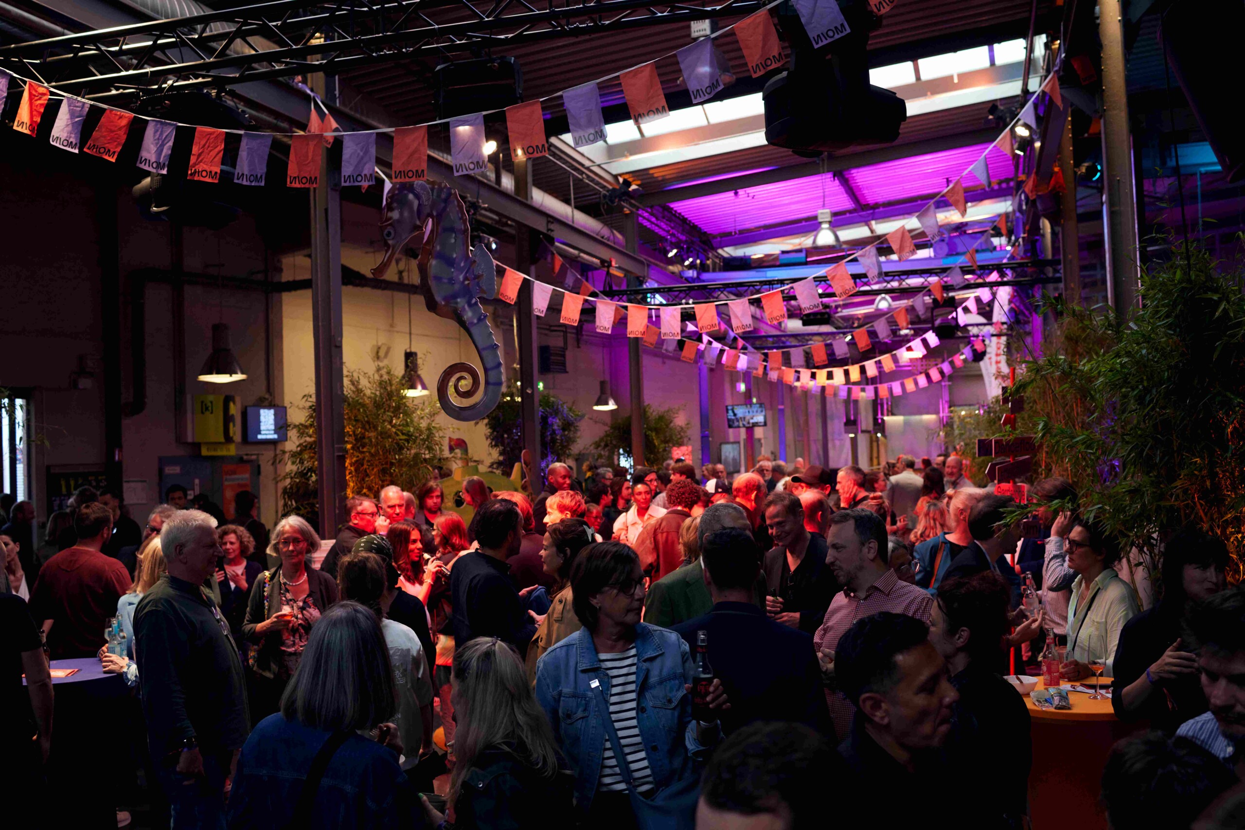 Menschenmenge feiert alljährliches Branchentreffen der MOIN Filmförderung in einer bunt beleuchteten Halle mit Seepferdchen-Dekoration und Girlanden.