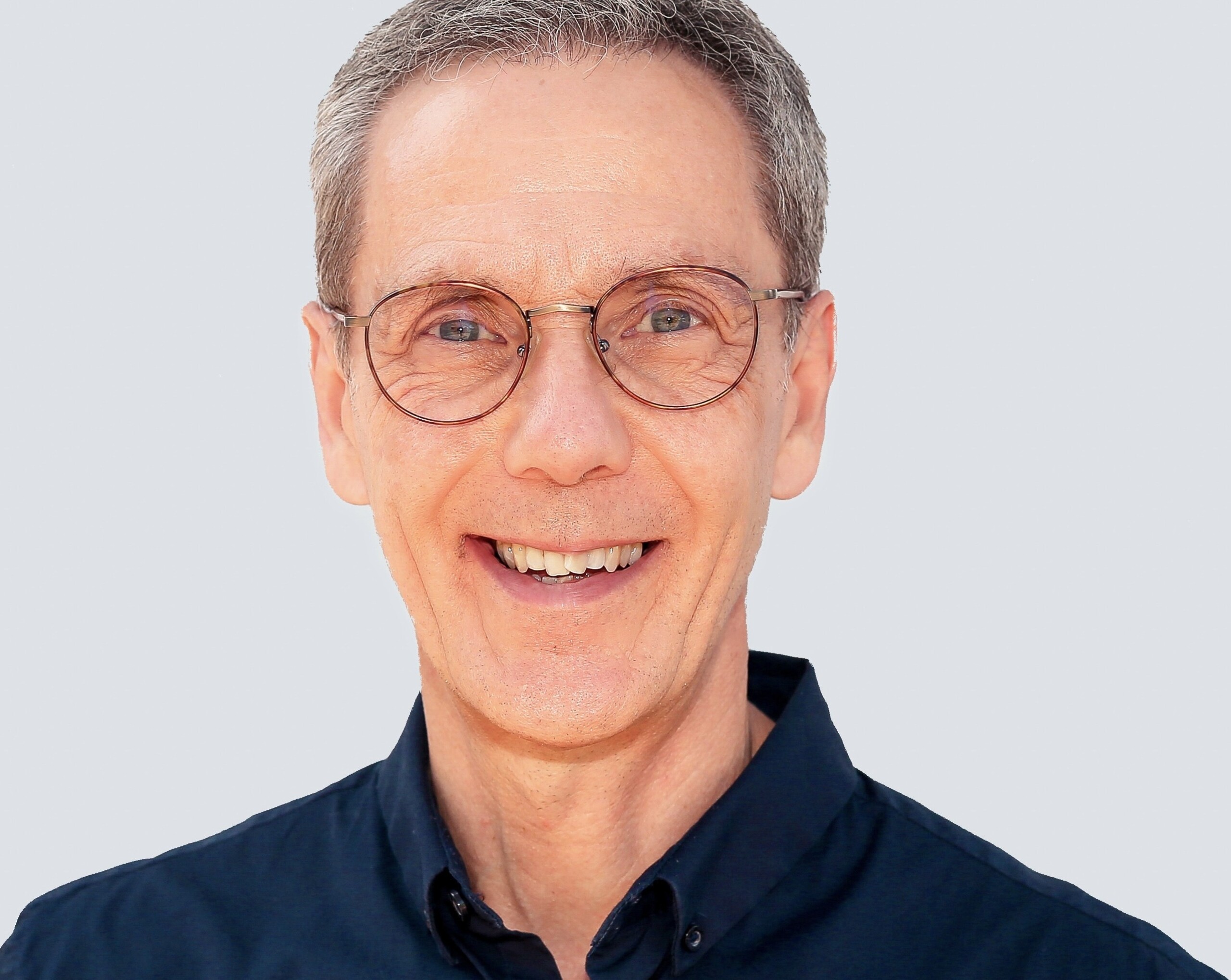 Smiling middle-aged man with short grey hair, round glasses, and a dark blue shirt against a plain background.