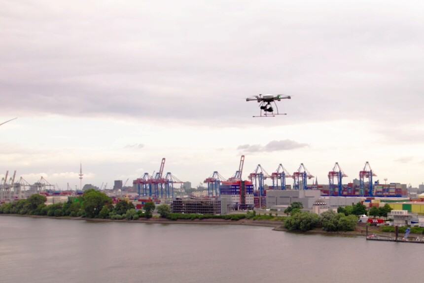 Drohne fliegt über Hafen mit Containerkränen und Gebäuden an einem bewölkten Tag.