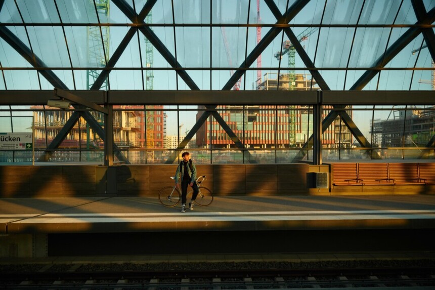 Person mit Fahrrad wartet auf Bahnsteig in moderner, glasüberdachter Station bei Sonnenuntergang.