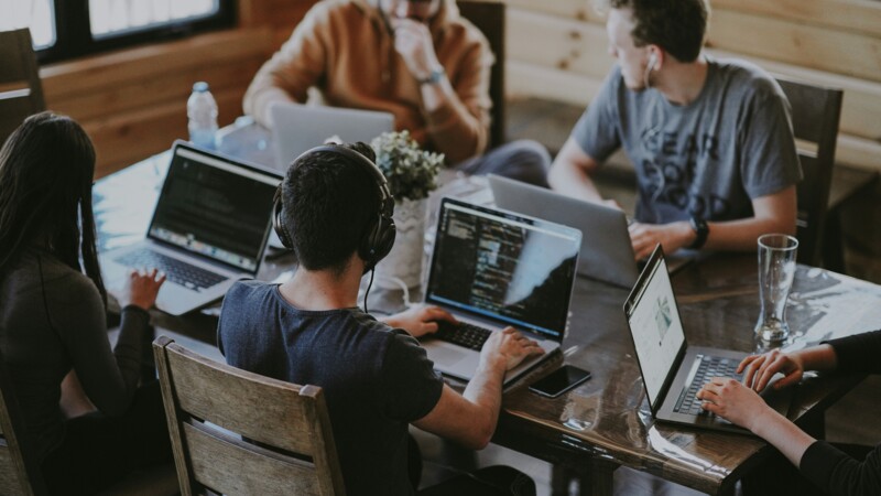 Junge Menschen sitzen mit Laptops um einem Schreibtisch in einem Großraumbüro