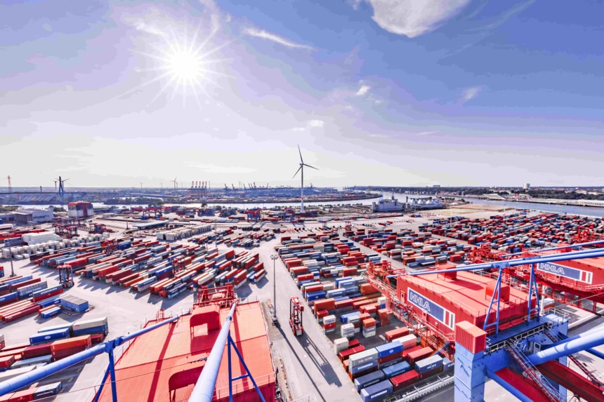 Luftaufnahme eines großen Containerhafens mit zahlreichen Containern, Kränen und einer Windkraftanlage bei Sonnenschein.