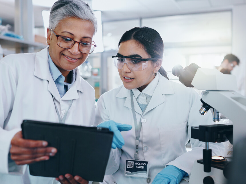 Zwei Wissenschaftlerinnen im Labor besprechen Daten auf einem Tablet neben einem Mikroskop.