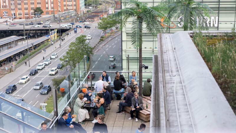 Menschen sitzen auf der Dachterasse des Jupiter mit Blick auf eine belebte Straße und den Hamburger Hauptbahnhof. 