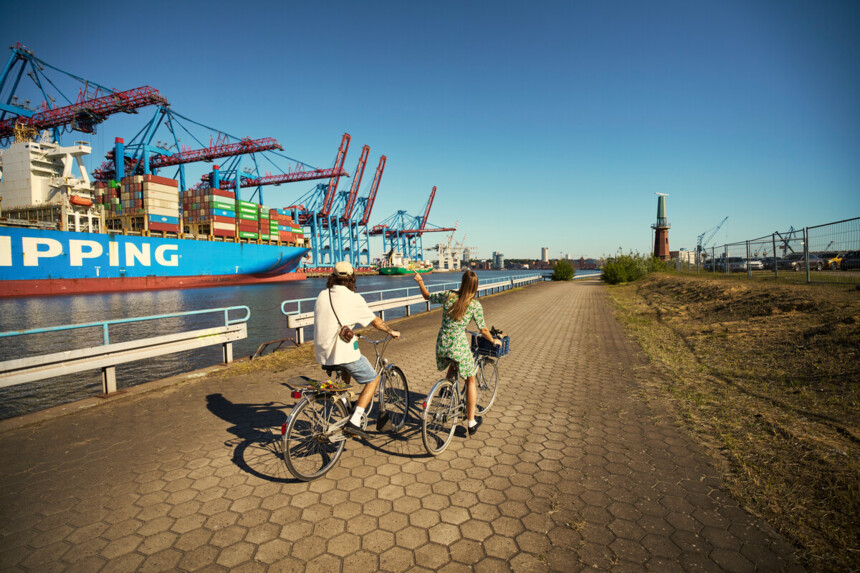 Zwei Personen fahren mit Fahrrädern entlang eines Hafens mit Containerschiffen und Kränen bei sonnigem Wetter.