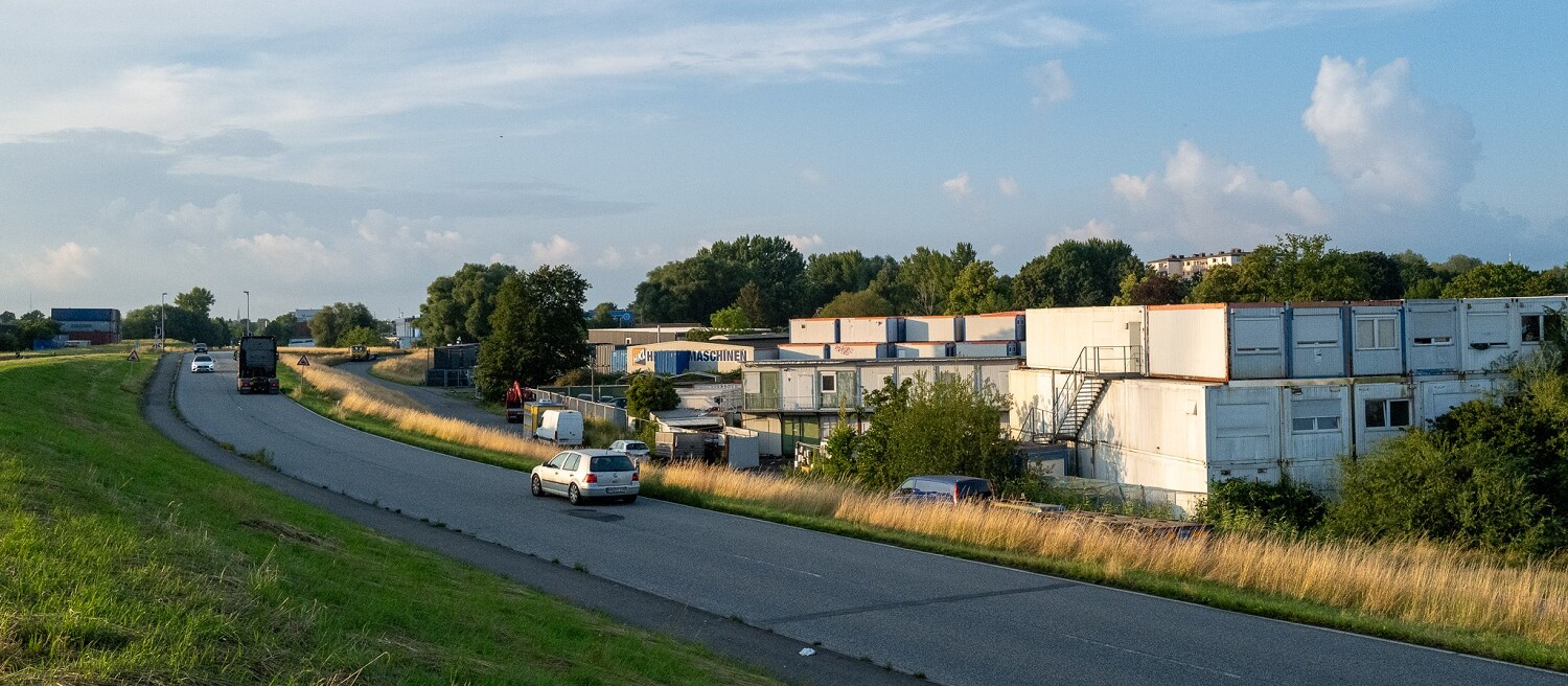 Street with stacked conatiners on one side and green spaces on the other.