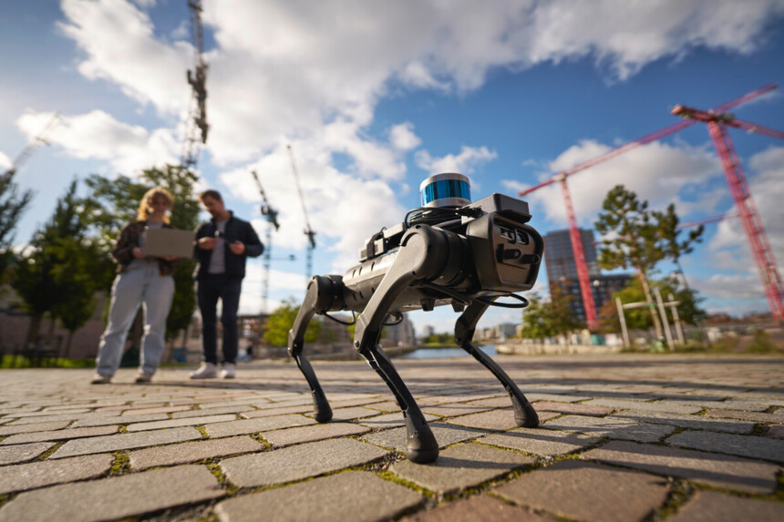 Vierbeiniger Roboter auf Pflastersteinweg mit zwei Personen und Baukränen im Hintergrund bei sonnigem Wetter.