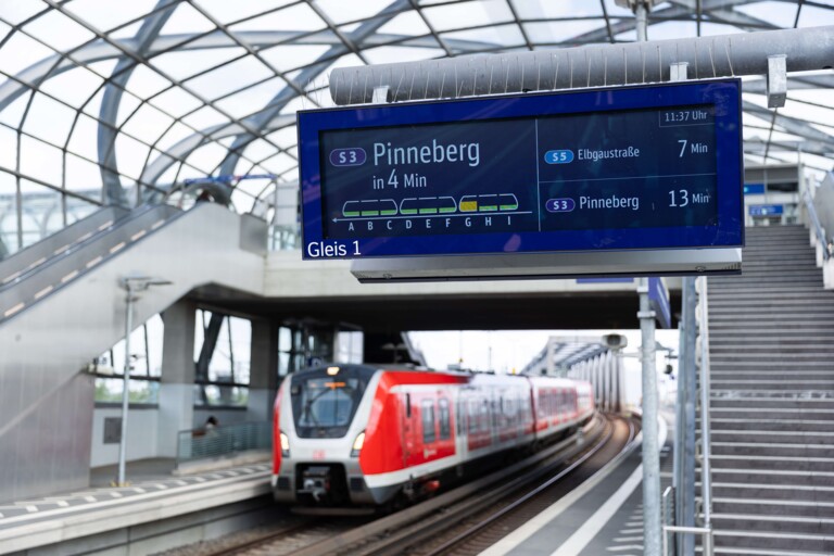 Elektronische Anzeigetafel am Bahnsteig mit Zugankünften und eine einfahrende rote S-Bahn