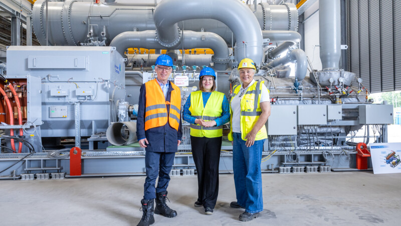 Three people with helmets and high-vis vests in front of an industrial plant 