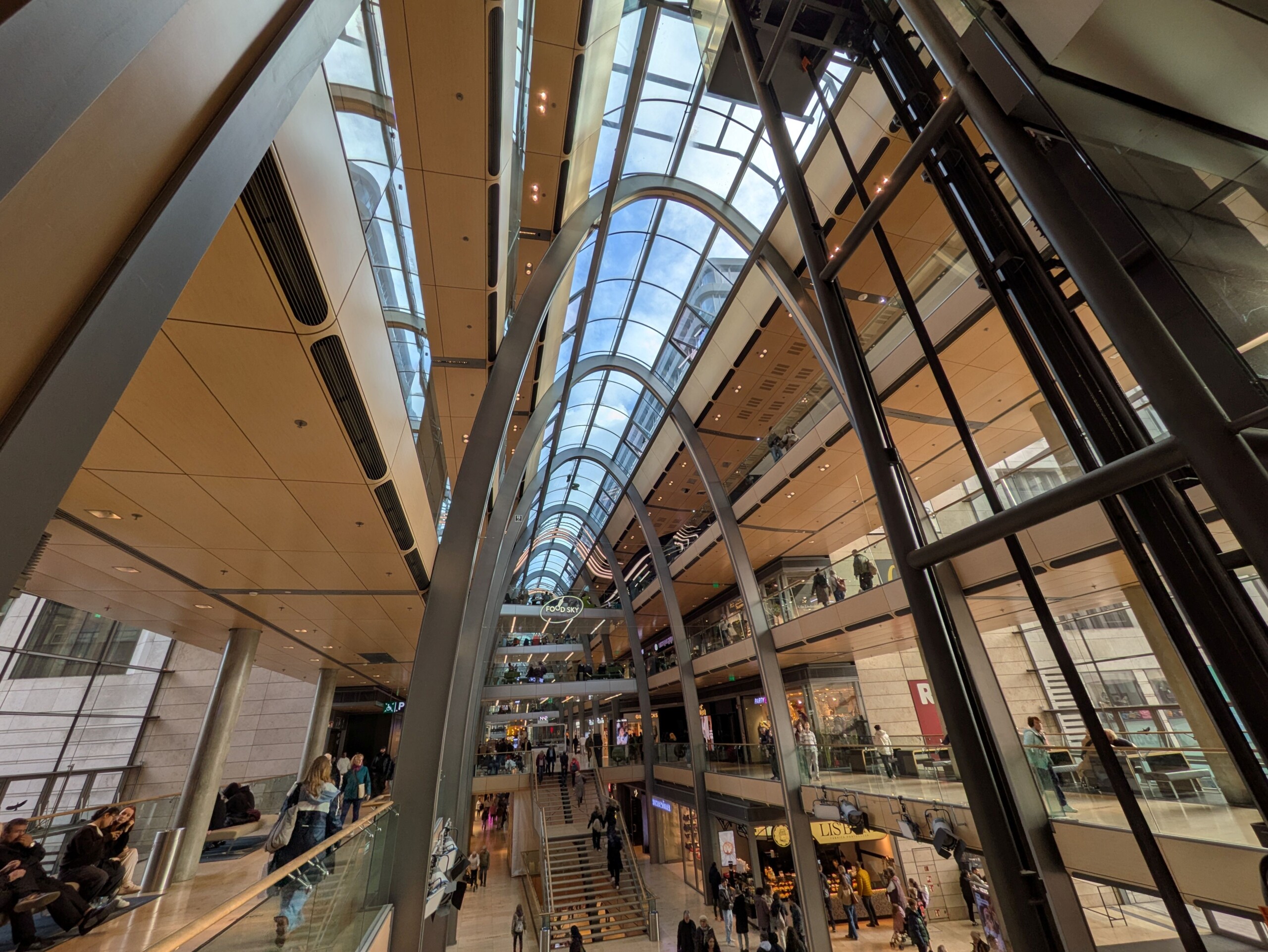 Innenansicht eines modernen Einkaufszentrums mit Glasdach und mehreren Etagen voller Besucher.