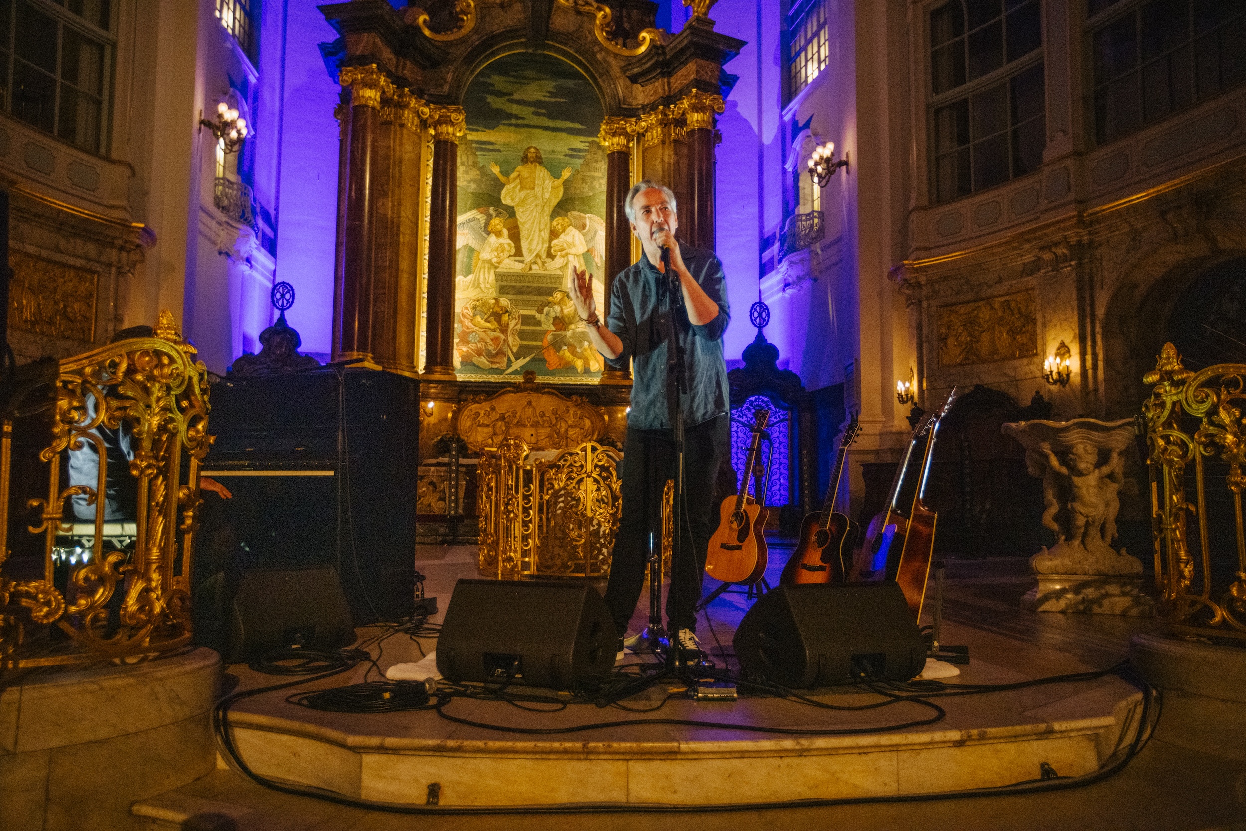 Mann singt mit Mikrofon auf Bühne in einer Kirche mit Gitarren und barockem Altar im Hintergrund.