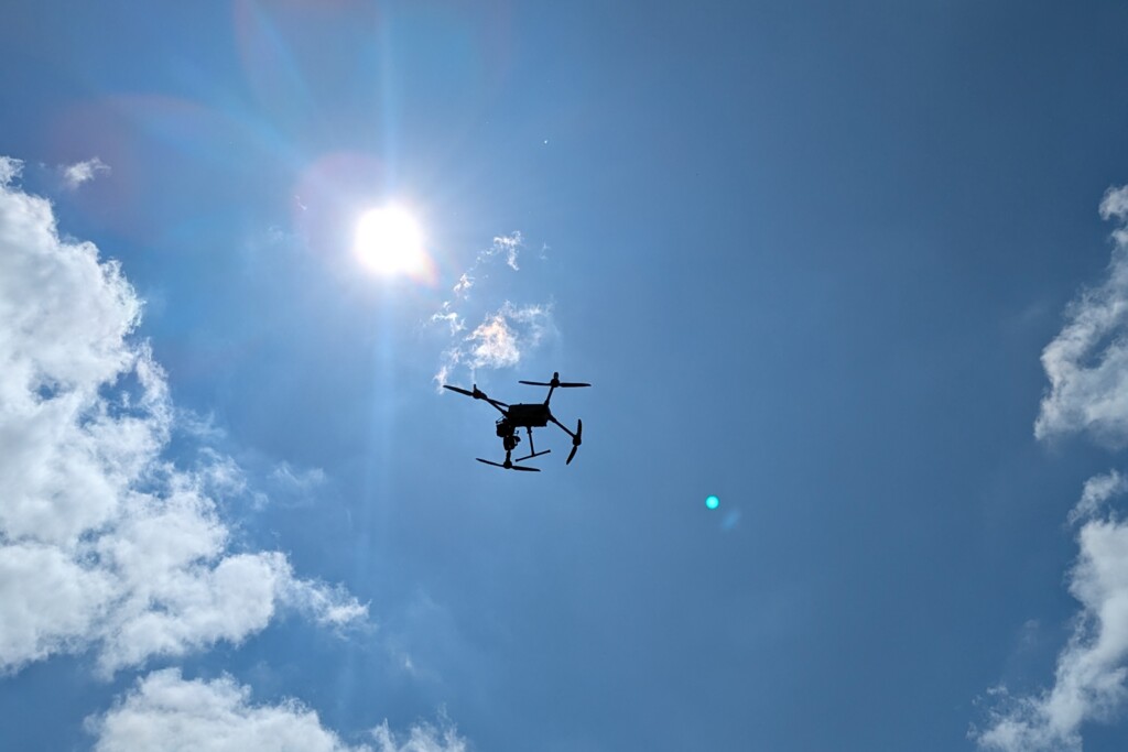 Drohne fliegt vor strahlend blauem Himmel mit Sonne und vereinzelten Wolken.