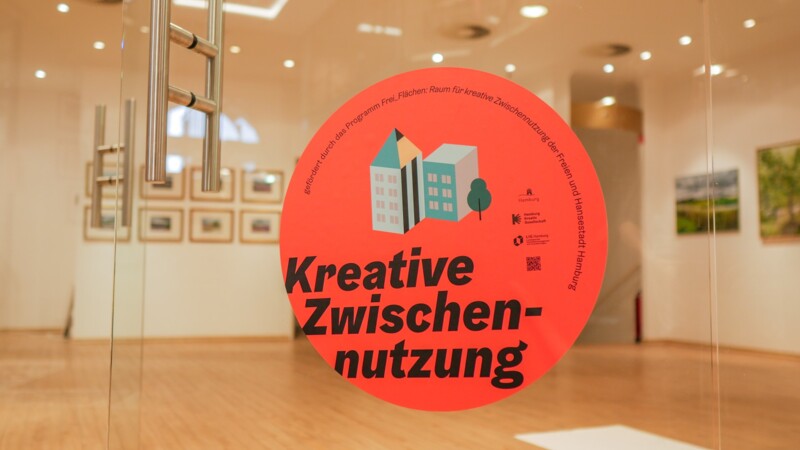 Kreative Zwischennutzung Symbolbild: Eine Glastür zu einem hellen Raum mit Kuns an den Wänden, auf der Glastür klebt ein runder Aufkleber, auf dem "Kreative Zwischennutzung" steht