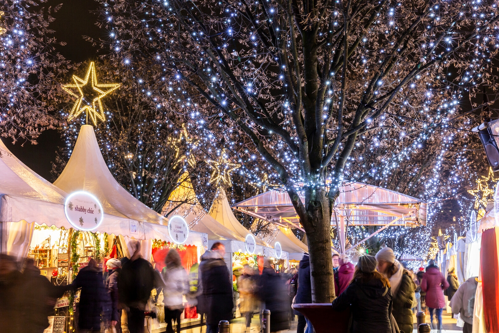 Weihnachtsmarkt bei Nacht mit beleuchteten Ständen, Sternenlichtern und festlich geschmückten Bäumen.