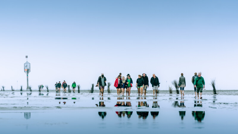 Gruppe von Menschen wandert bei Ebbe barfuß durch das Watt mit Spiegelungen im Wasser.
