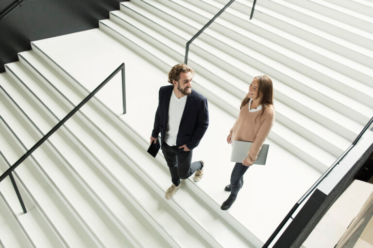 Zwei junge Erwachsene gehen lachend eine breite weiße Treppe hinauf, einer mit Tablet, die andere mit Laptop.