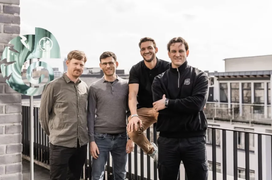 Vier Männer stehen lächelnd auf einer Dachterrasse mit Stadtblick und einem grünen Kunstwerk im Hintergrund.