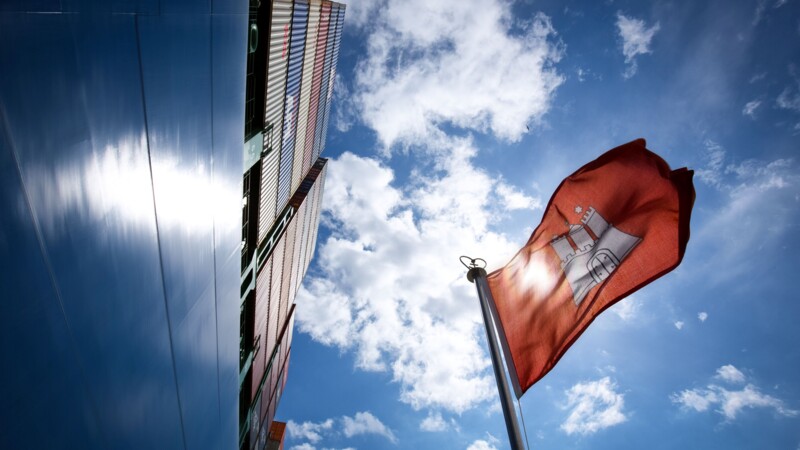 Blick nach oben auf Container-Schiff mit Hamburg-Flagge vor blauem Himmel und Wolken.