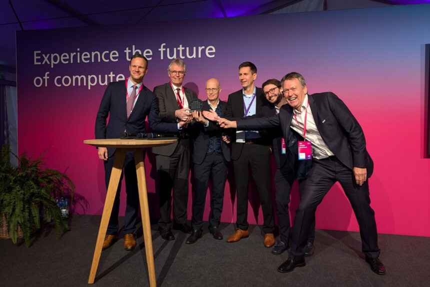 Sechs Männer in Anzügen präsentieren gemeinsam ein rundes, technisches Gerät vor einem „Experience the future of compute“-Hintergrund.