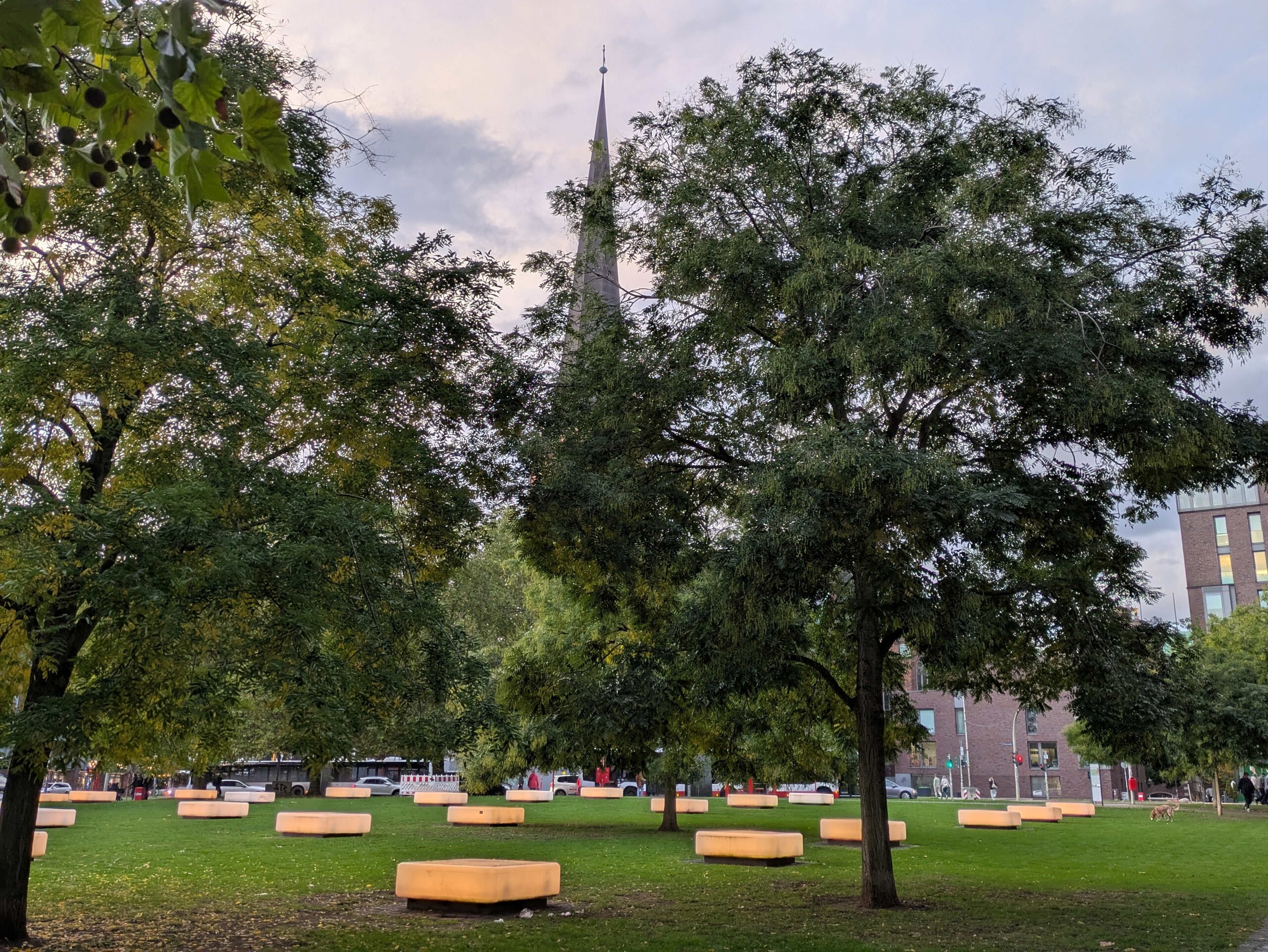 Grüner Park mit leuchtenden Sitzbänken und Kirchturmspitze im Hintergrund bei bewölktem Himmel.