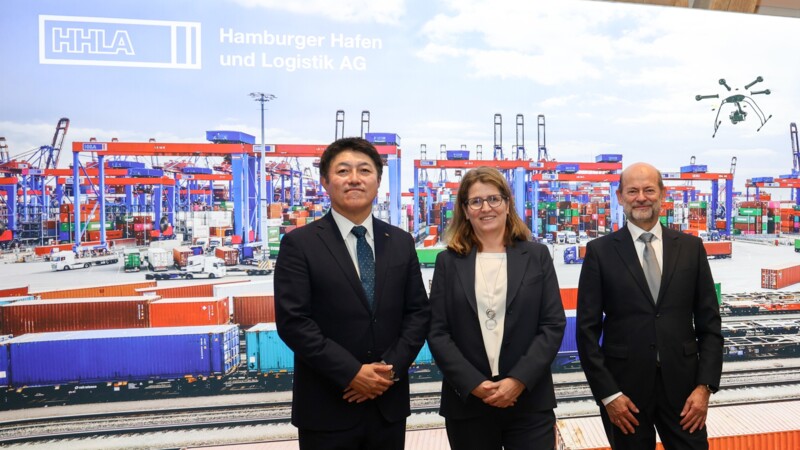 Drei Personen in Anzügen vor einem Hintergrund mit Containerhafen und HHLA-Logo.