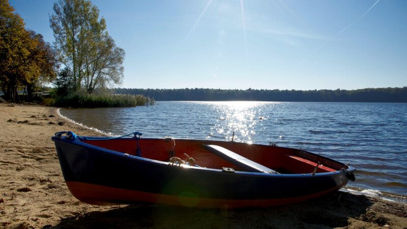 Ruderboot am sandigen Ufer eines sonnigen Sees mit Bäumen im Hintergrund.