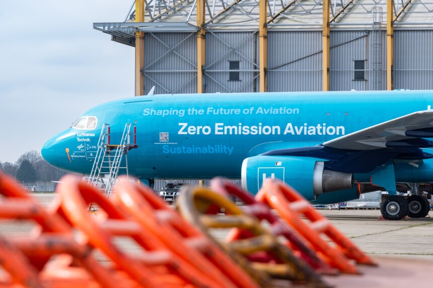 Blaues Flugzeug mit Aufschrift „Zero Emission Aviation“ auf dem Rollfeld vor Hangar.