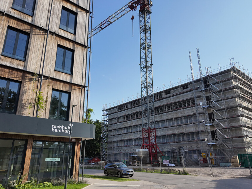 Baukran und Baustelle neben modernem Holzgebäude mit Schild „techhub hamburg“ bei klarem Himmel.