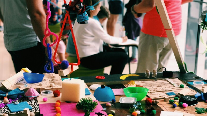 MakerLab Symbolbild: Bunte Bastelarbeiten auf einem Tisch, im Hintergrund Menschen, die an etwas tüfteln