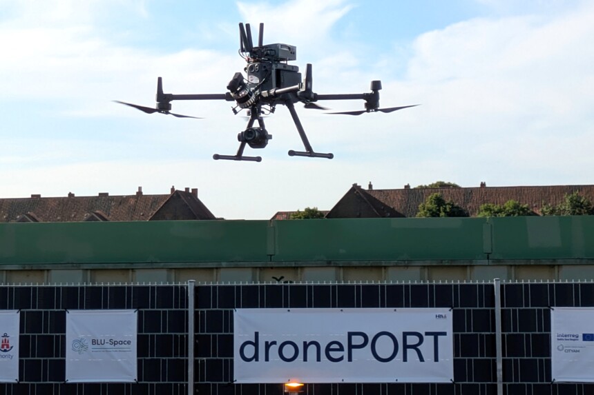 Schwarze Drohne mit Kamera fliegt vor Gebäuden über einem Zaun mit Banner „dronePORT“.