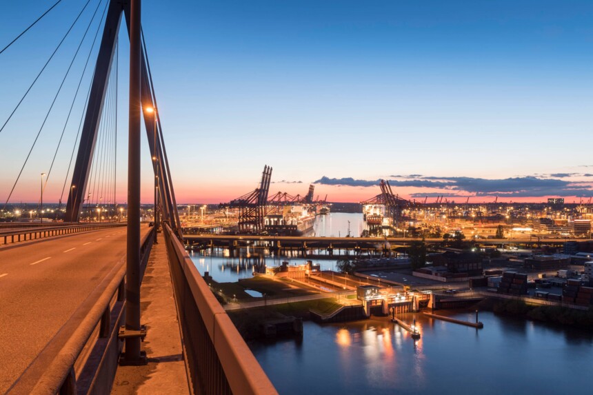 Blick von einer beleuchteten Brücke auf einen Hafen mit Kränen und Containerschiffen bei Sonnenuntergang.