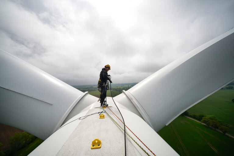 Techniker sichert sich auf einem Windrad zur Wartung bei bewölktem Himmel und grünem Landschaftshintergrund.