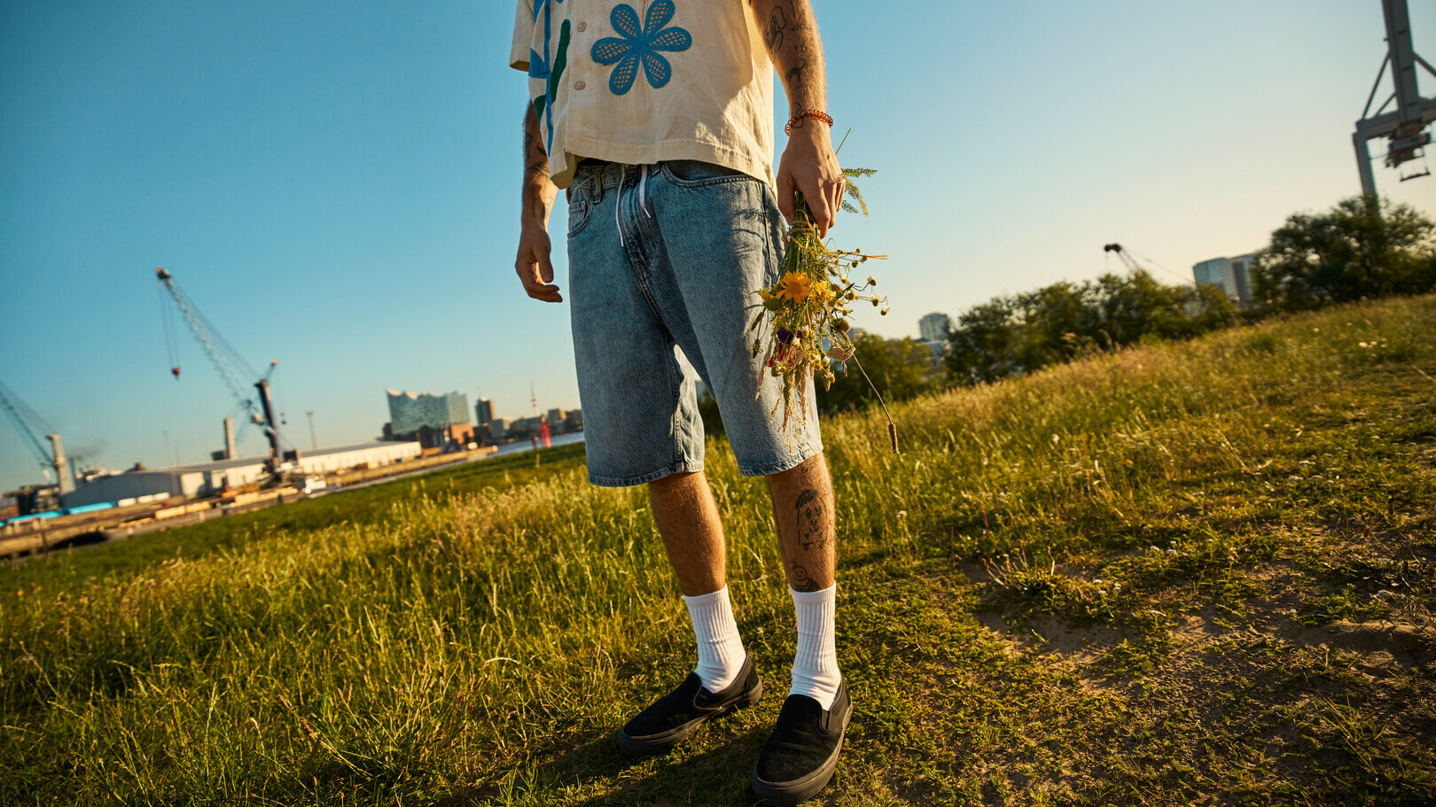 Person mit Tattoos, Jeansshorts und Blumen in der Hand steht auf einer Wiese bei Sonnenuntergang, im Hintergrund der Hamburger Hafen.