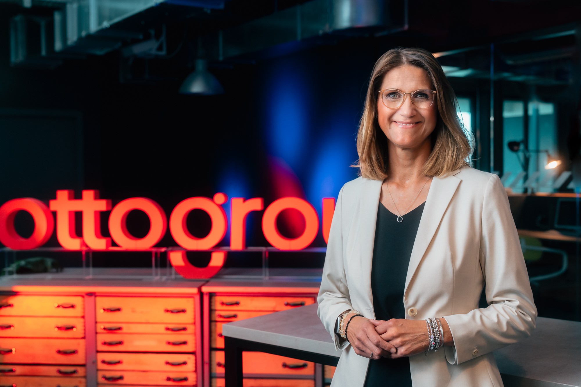 Portrait photo of Katy Roewer, member of the Otto Group Executive Board. She is wearing a light-colored blazer, a black T-shirt, glasses and has blonde hair. In the background you can see the Group name "ottogroup" in illuminated lettering