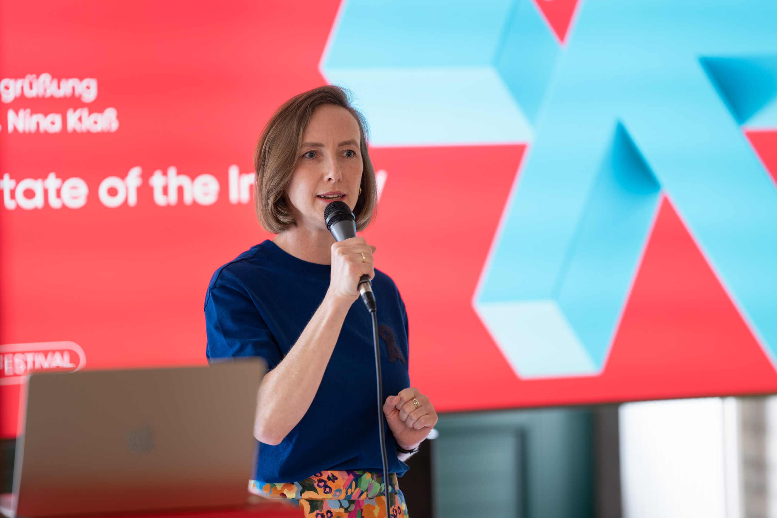 Nina Klaß, Leiterin von Next Media bei ihrer Eröffnungsrede. Sie trägt ein T-Shirt und eine gemusterte Hose und hält ein Mikrofon
