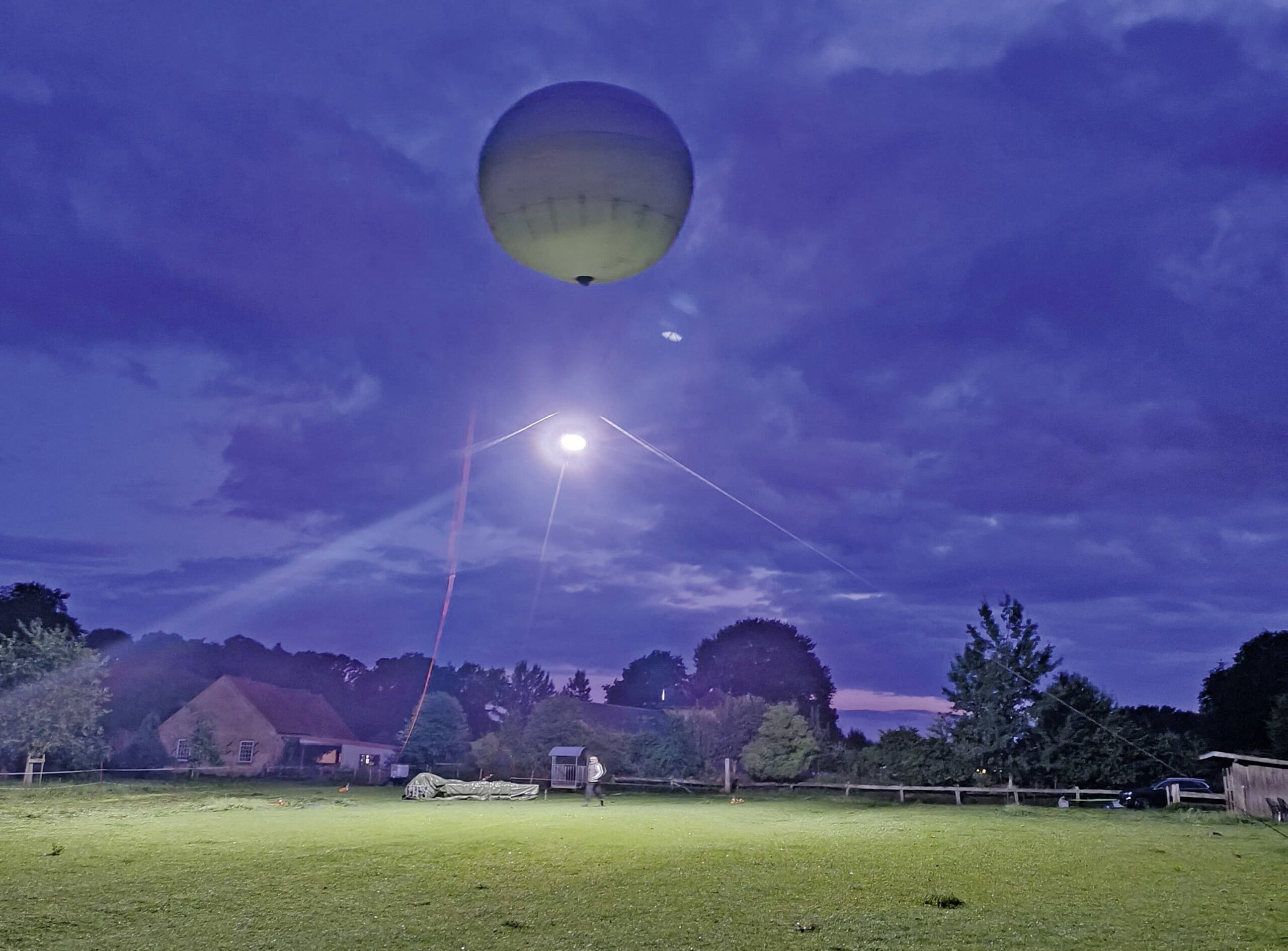Humanilog-Ballon bei Dämmerung über einem Feld mit Haus und Bäumen im Hintergrund.