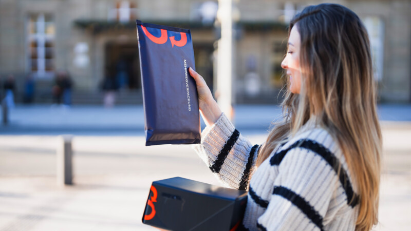 Frau hält dunkelblaue Mehrwegverpackungen mit roter Schrift im Freien.