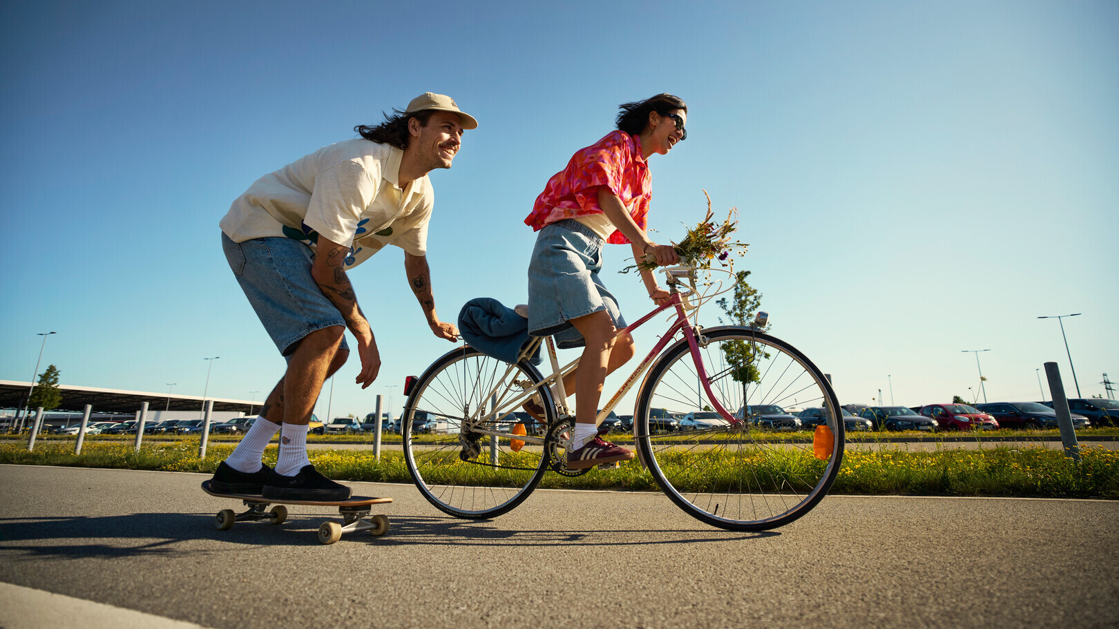 Junge Frau fährt Fahrrad mit Blumen im Korb, während junger Mann auf Skateboard hinterherfährt.