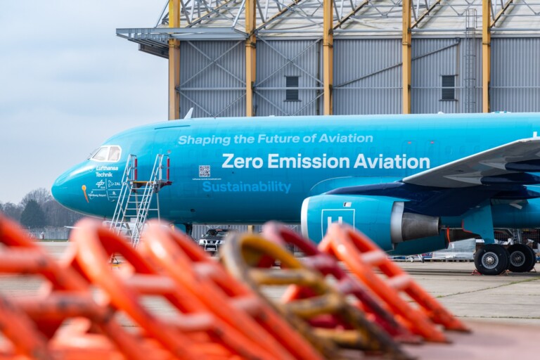 Blaues Flugzeug mit der Aufschrift „Zero Emission Aviation“ vor Hangar und orangefarbenen Absperrungen.