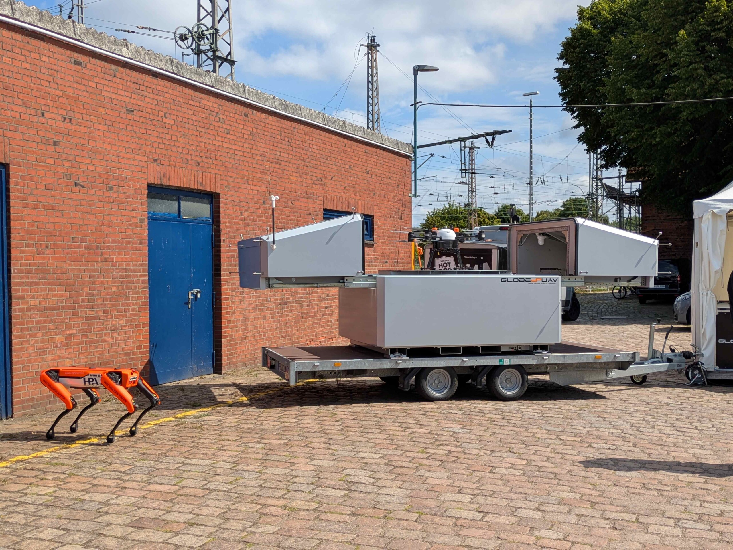 Two orange robotic dogs next to a trailer-mounted drone station outside a brick building.