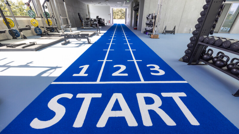 Innenraum eines Fitnessstudios mit blauer Laufbahn und verschiedenen Trainingsgeräten.