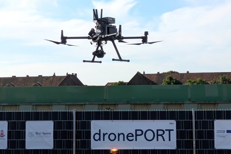 Schwarze Drohne mit Kamera fliegt über einen Zaun mit Bannern des Projekts dronePORT.