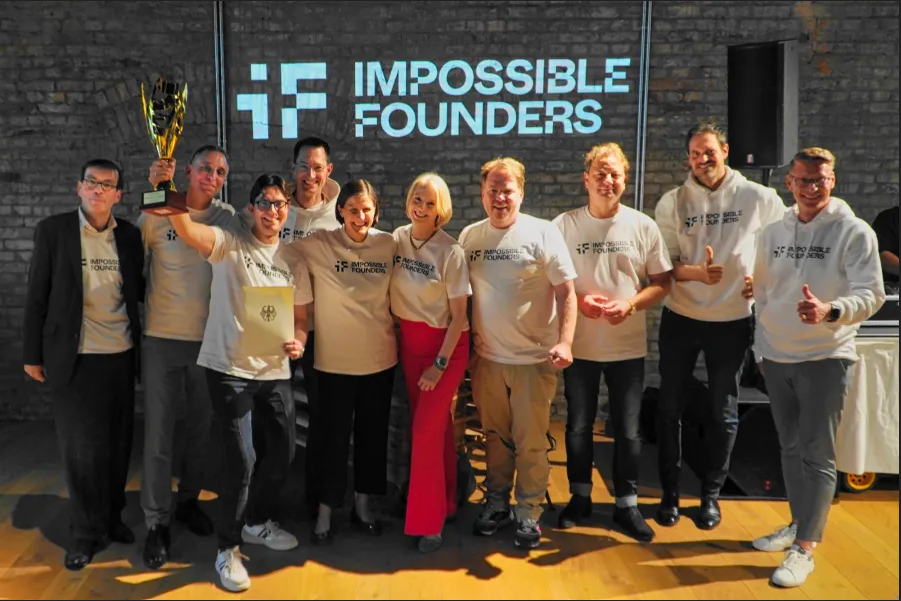 Group of people wearing Impossible Founders shirts celebrating with a trophy in front of a branded wall.
