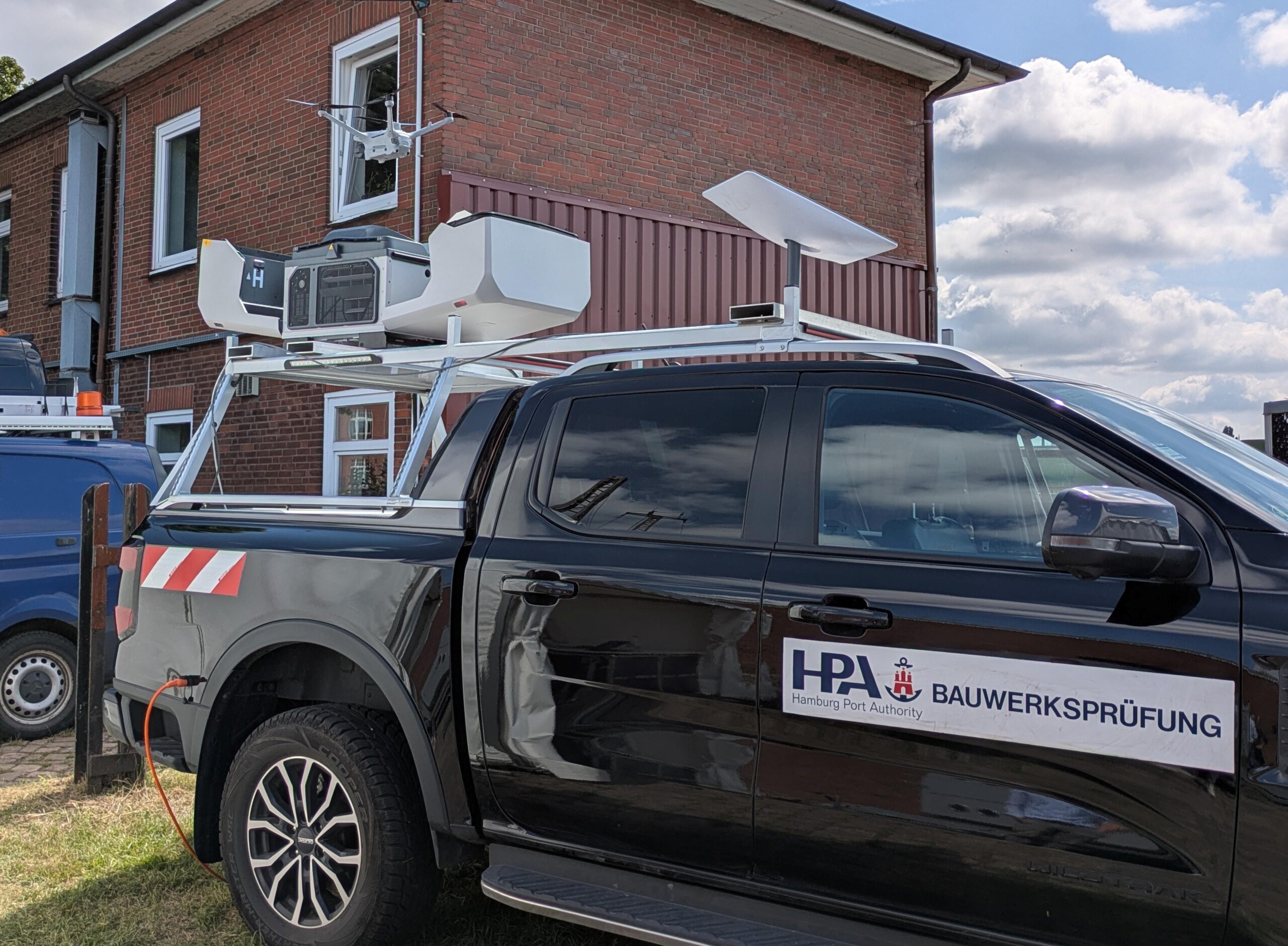 Black Hamburg Port Authority vehicle with structural inspection equipment mounted on top, parked beside a brick building.