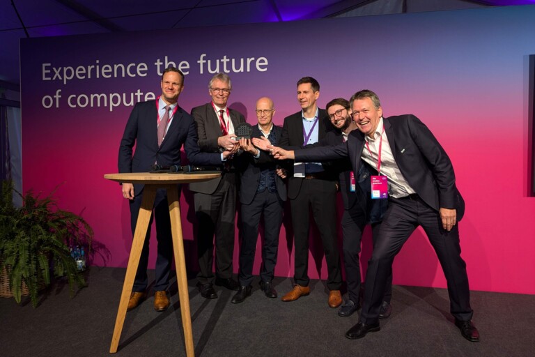 Sechs Männer in Anzügen präsentieren gemeinsam ein technisches Gerät vor einem Banner mit dem Text „Experience the future of compute“.